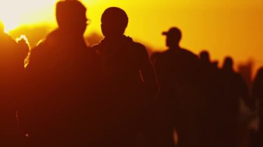 Bir grup insan, arka planda güneş batarken şehir caddesinde yürür ve sıcak, parlak bir atmosfer yaratır..