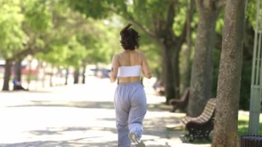 Bir kadın parkta patikadan aşağı koşuyor. Beyaz bir atlet ve gri pantolon giyiyor.