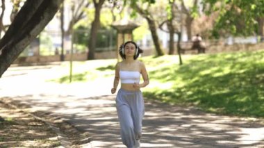 Bir kadın parkta bir yolda koşuyor. Beyaz bir atlet ve gri pantolon giyiyor.