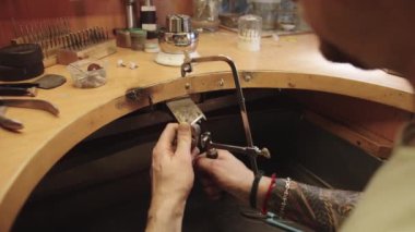 Bir adam bir mücevher üzerinde çalışıyor, bir metal parçasını kesmek için bir alet kullanıyor. Sahne, erkeğin ellerine ve kullandığı alete odaklanmış durumda. Başka nesne ya da insan yok.