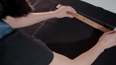 Bir kadın bir cetvel tutuyor ve bir kumaş parçasını işaretliyor. Cetvel sayılarla işaretlenmiş ve kişi kumaşa çizgi çekmek için kalem kullanıyor.