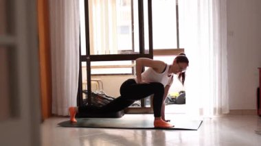 Bir kadın oturma odasında yoga pozu veriyor. Beyaz atlet ve siyah pantolon giyiyor.