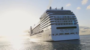 MS Orkestrası adında büyük bir yolcu gemisi okyanusa yelken açıyor. Gemi beyaz ve yan tarafında mavi bir şerit var. Güneş arka planda batıyor, sıcak ve huzurlu bir atmosfer yaratıyor.