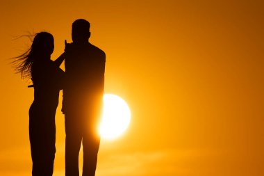 Güneşin önünde kadının erkeğin koluna dokunduğu bir çift. Sahne romantik ve samimi.