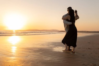 Gün batımında sahilde dans eden bir çift. Adam, kadının kolunu tutuyor. Sahne romantik ve huzurlu.