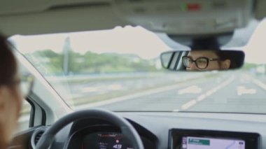Bir kadın araba kullanıyor ve dikiz aynasından yansımasına bakıyor. Gülümsüyor, bu da araba sürmekten hoşlandığını gösteriyor.