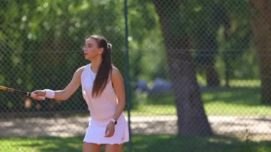 Arka planda çitlerle kortta tenis oynayan bir kadın. Beyaz bir gömlek ve beyaz bir etek giyiyor.