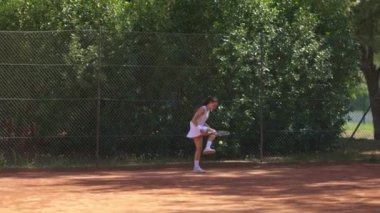 Elinde tenis raketiyle tenis kortunda yürüyen bir kadın. Saray ağaçlar ve çitlerle çevrili.