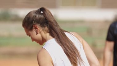 Uzun saçlı bir kadın pis bir tarlada dururken gülümsüyor. Beyaz bir atlet giyiyor ve keyfine bakıyor.