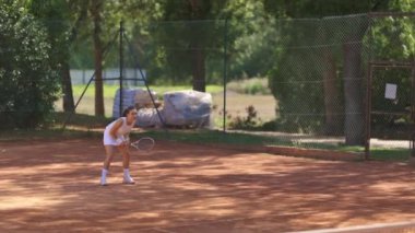 Bir kadın kortta tenis oynuyor. Mahkeme bir çitle çevrili ve çitin yanında bir çanta yığını var.