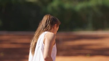 Beyaz atletli bir kadın tenis kortunda yürüyor. Aşağıya, yere bakıyor.