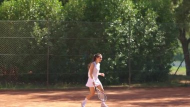 Bir kadın kortta tenis oynuyor. Beyaz bir elbise giyiyor ve elinde bir tenis raketi tutuyor.