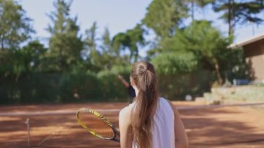 Uzun saçlı bir kadın tenis kortunda yürüyor. Elinde bir tenis raketi tutuyor ve oyuna odaklanmış durumda.