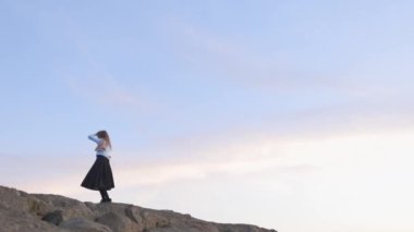 Kayalık bir yamaçta mavi gömlek ve siyah etek giyen bir kadın var. Elinde bir eşarp tutuyor ve manzaranın tadını çıkarıyor. Gökyüzü açık ve mavi