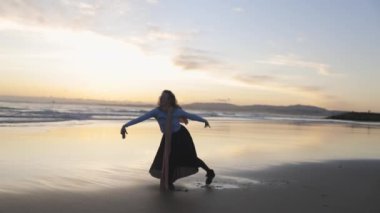 Bir kadın sahilde dans ediyor. Gökyüzü turuncu ve su sakin. Kadın siyah etek ve beyaz gömlek giyiyor.