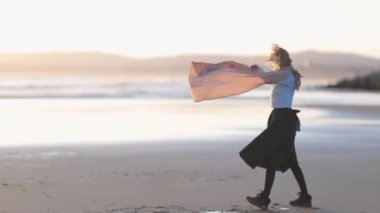 Bir kadın sahilde boynunda eşarpla dans ediyor. Sahne sakin ve huzurlu, arkada okyanus var. Kadınların hareketleri zarif ve akıcıdır.