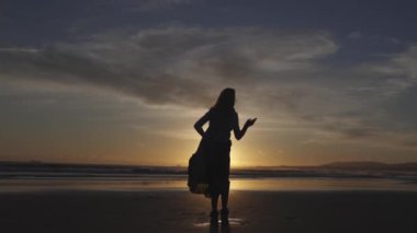 Gün batımında bir kadın sahilde yürüyor. Gökyüzü bulutlarla dolu ve güneş uzaklardan batıyor. Kadın bir elbise giyiyor ve güzel manzaranın tadını çıkarıyor.