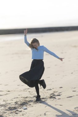 Bir kadın sahilde dans ediyor. O, mavi bir gömlek ve siyah bir etek giyiyor. Kumsal kumlu ve gökyüzü temiz.
