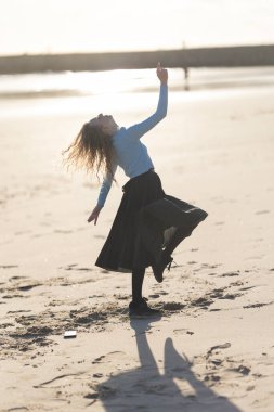 Bir kadın kumsalda kollarını açmış dans ediyor. Kumsal boş ve güneş parlıyor.