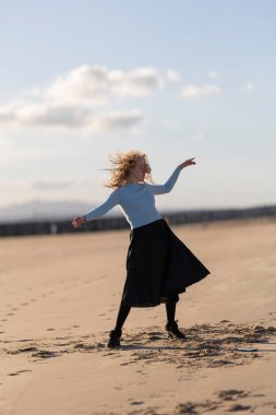 Plajda siyah etekli ve mavi gömlekli bir kadın duruyor. Dans ediyor ve eğleniyor. Plaj manzarası ve kadınların tasasız tavırları bir rahatlama hissi yaratıyor.