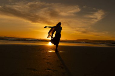 Sahilde bir kadın duruyor, arka planda güneş batıyor. O bir eşarp takıyor ve dans ediyor. Kadın günbatımının güzelliğinin tadını çıkarırken sahne huzurlu ve huzurlu.