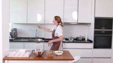 Bir kadın elinde beyaz bir tezgah ve kahverengi bir önlükle mutfağa giriyor. Mutfakta lavabo, fırın ve mikrodalga fırın var. Tezgahta birkaç kase, fincan ve kaşık var.