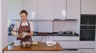 Bir kadın mutfakta beyaz bir tezgahla yemek pişiriyor. Önlük ve eldiven giyiyor.