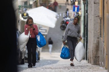 11 Aralık 2023, Lizbon, Portekiz - İki kadın sokakta yürüyor, biri cep telefonu tutuyor. Çanta taşıyorlar ve içlerinden biri yastık tutuyor.
