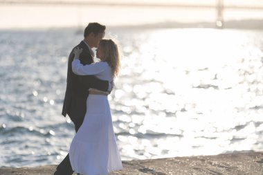 Arkalarında güneş batarken bir çift, kayalık bir sahilde dans eder ve suyun üzerinde sıcak bir parıltı yaratır..