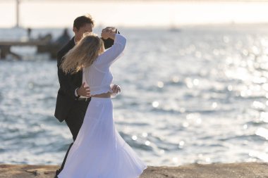 Bir kadın ve bir erkek gün batımında köpüklü bir denize bakan iskelede dans ediyorlar. Kadın dalgalı beyaz bir elbise giyer ve erkek koyu renk bir takım giyer..