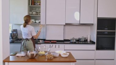 Bir kadın mutfak tezgahı ve bir sürü aleti olan bir mutfakta yemek pişiriyor. Beyaz önlük ve eldiven giyiyor.