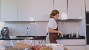 Bir kadın mutfakta yemek hazırlıyor. O bir önlük ve eldiven giyiyor. Mutfak temiz ve iyi düzenlenmiş.