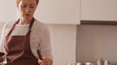 Bir kadın mutfakta yemek pişiriyor. O, beyaz bir gömlek ve önlük giyiyor. Elinde bir spatula ve kaşık tutuyor.