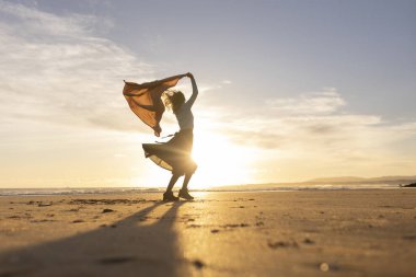 Bir kadın sahilde boynunda eşarpla dikiliyor. Gökyüzü güzel turuncu bir renktir ve güneş batıyor. Kadın anın ve güzel manzaranın tadını çıkarıyor.
