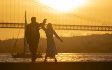 Bir çift su kenarında iskelede dans ediyor. Güneş arka planda batıyor, sıcak ve romantik bir atmosfer yaratıyor.
