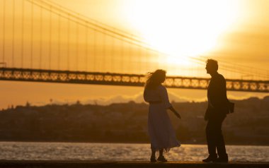 Bir çift, bir köprünün yakınındaki iskelede yürüyor. Arka planda güneş batıyor. Sahne romantik ve huzurlu.