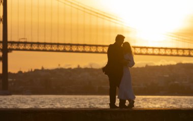 Arka planda güneş batarken bir çift köprünün önünde öpüşüyor. Sahne romantik ve samimi.