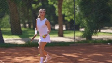 Bir kadın kortta tenis oynuyor. Beyaz bir elbise ve beyaz bir bluz giyiyor.