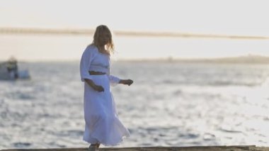 Beyaz elbiseli bir kadın suya bakan bir iskelede dans ediyor. Manzara sakin ve huzurlu, kadın elbisesi rüzgarda dalgalanırken. Su sakin.