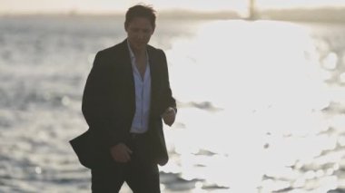 Takım elbiseli bir adam sahilde yürüyor. Su sakin ve güneş batıyor.