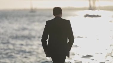 Takım elbiseli bir adam sahilde dans ediyor. Su sakin ve gökyüzü kapalı.