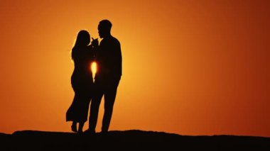 Bir çift gün batımına karşı siluetlendi, kadının elini tutan adamla. Sahne romantik ve samimi, çift birlikte duruyor ve güzel gün batımını seyrediyor.