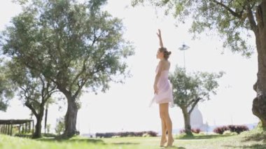 Pembe elbiseli bir kadın ağaçlarla çevrili bir parkta dans ediyor. Güzel havanın ve yeşil çimlerin tadını çıkarıyor..