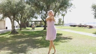 Pembe elbiseli genç bir kadın güneşli bir günde parkta dans eder. Gülümsüyor ve dans ederken kollarını zarifçe hareket ettiriyor..