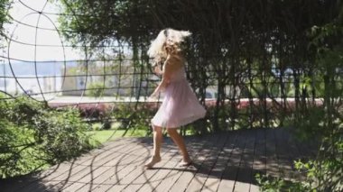 Pembe elbiseli genç bir kadın yemyeşil bir araziyle çevrili bir parkta ahşap bir platformda dans ediyor. Güneş parlıyor, sahne boyunca gölgeler dökülüyor..