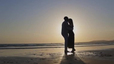 Romantik bir kumsalda gün batımının tadını çıkarıyorlar. Silüet bir öpücük paylaşıyorlar. Huzurlu bir ortamda sevgi ve sevgi uyandırıyorlar.