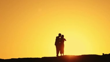 Gün batımında kucaklaşan bir çiftin silueti, altın gökyüzü ile romantik ve huzurlu bir atmosfer yaratıyor..