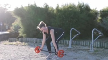 Parkta elinde kırmızı bir topla bir kadın duruyor. Park ağaçlarla çevrili ve arka planda bir bank var.