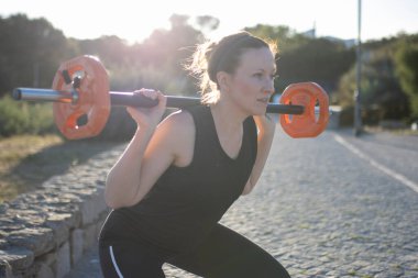 Bir kadın ağır bir barı kaldırıyor. Siyah bir atlet ve siyah pantolon giyiyor.