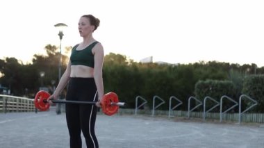 Bir kadın kırmızı ve siyah bir ağırlığı kaldırıyor. Kadın gülümsüyor ve antrenmanından zevk alıyor.